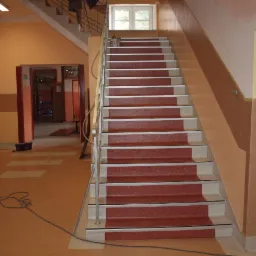 Klatka schodowa w budynku użyteczności publicznej z nowo zamontowanymi, czerwonymi nakładkami dywanowymi na stopniach, zabezpieczonymi aluminiowymi listwami. Widoczne ślady po kleju na podłodze.