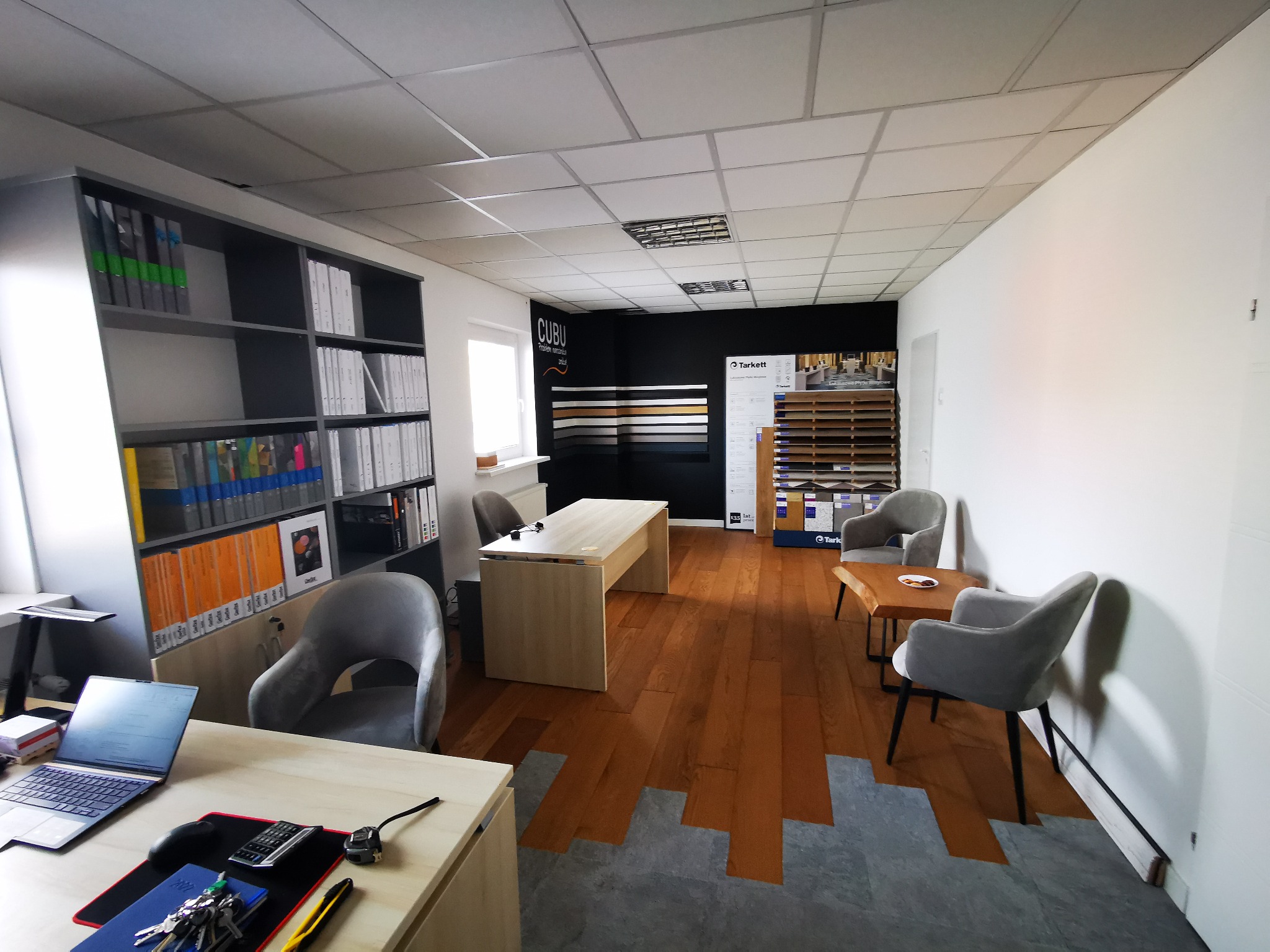 Biuro z ekspozycją podłóg: deska warstwowa w odcieniach brązu łączy się z szarą płytką dywanową. W tle widoczne wzorniki podłóg Tarkett i Cubu.