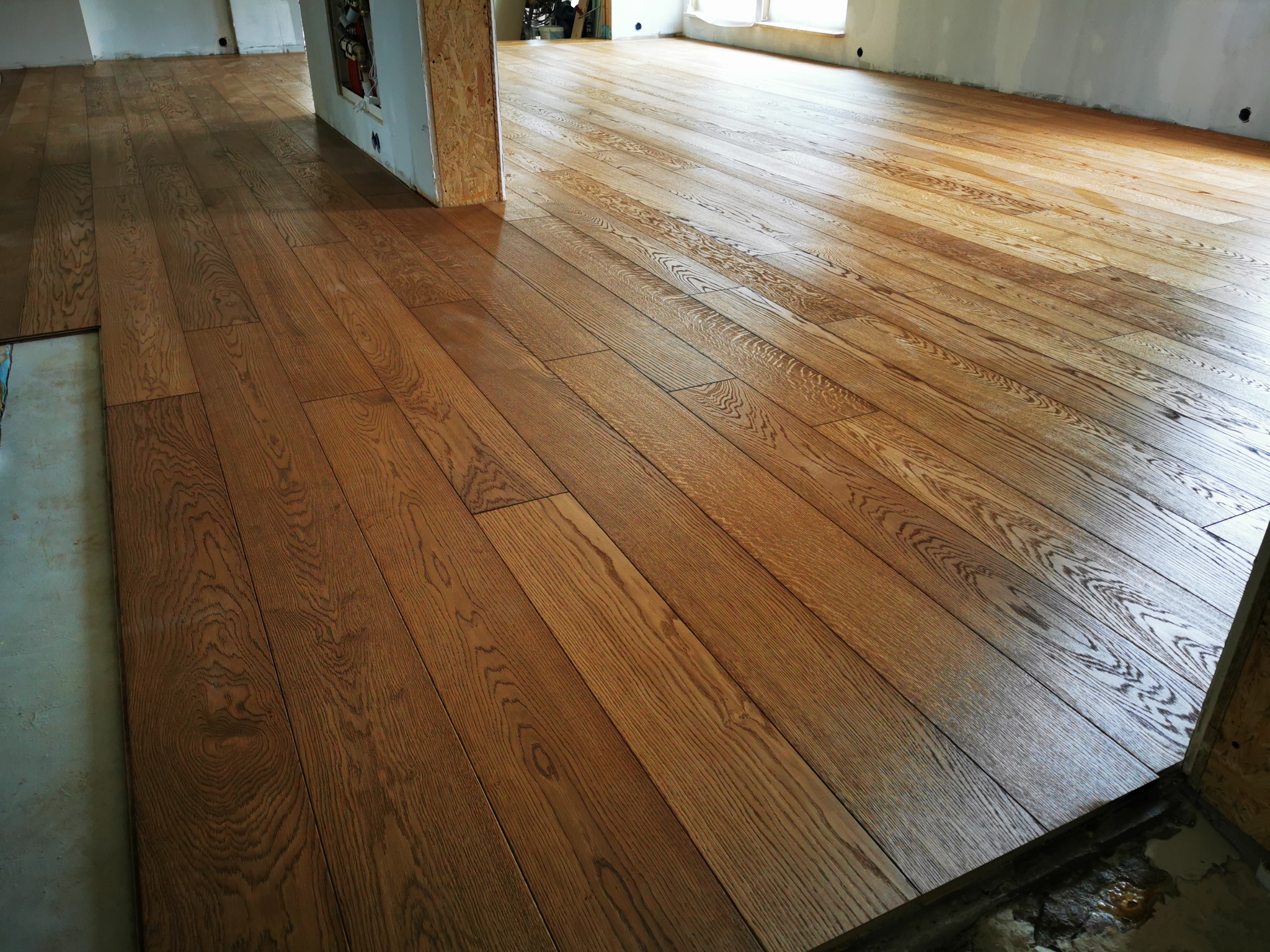 Deski podłogowe z wyraźnym usłojeniem, ułożone w nowym pomieszczeniu, widoczne surowe ściany i resztki materiałów budowlanych.