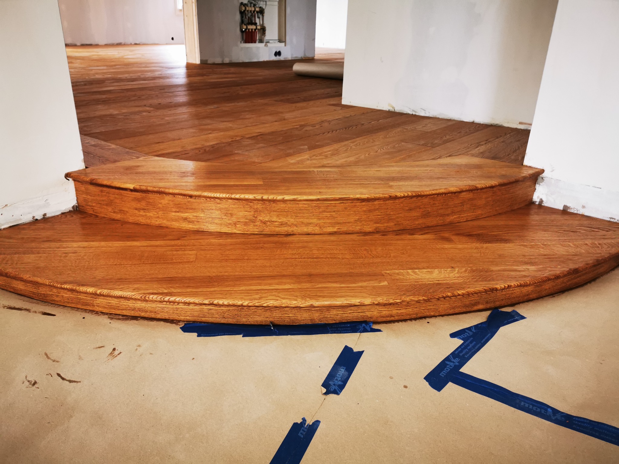 Dwu-stopniowe schody z drewna dębowego, z widocznym parkietem ułożonym w jodełkę francuską, zabezpieczone taśmą malarską na papierze ochronnym podłogi, w tle widoczne narzędzia.