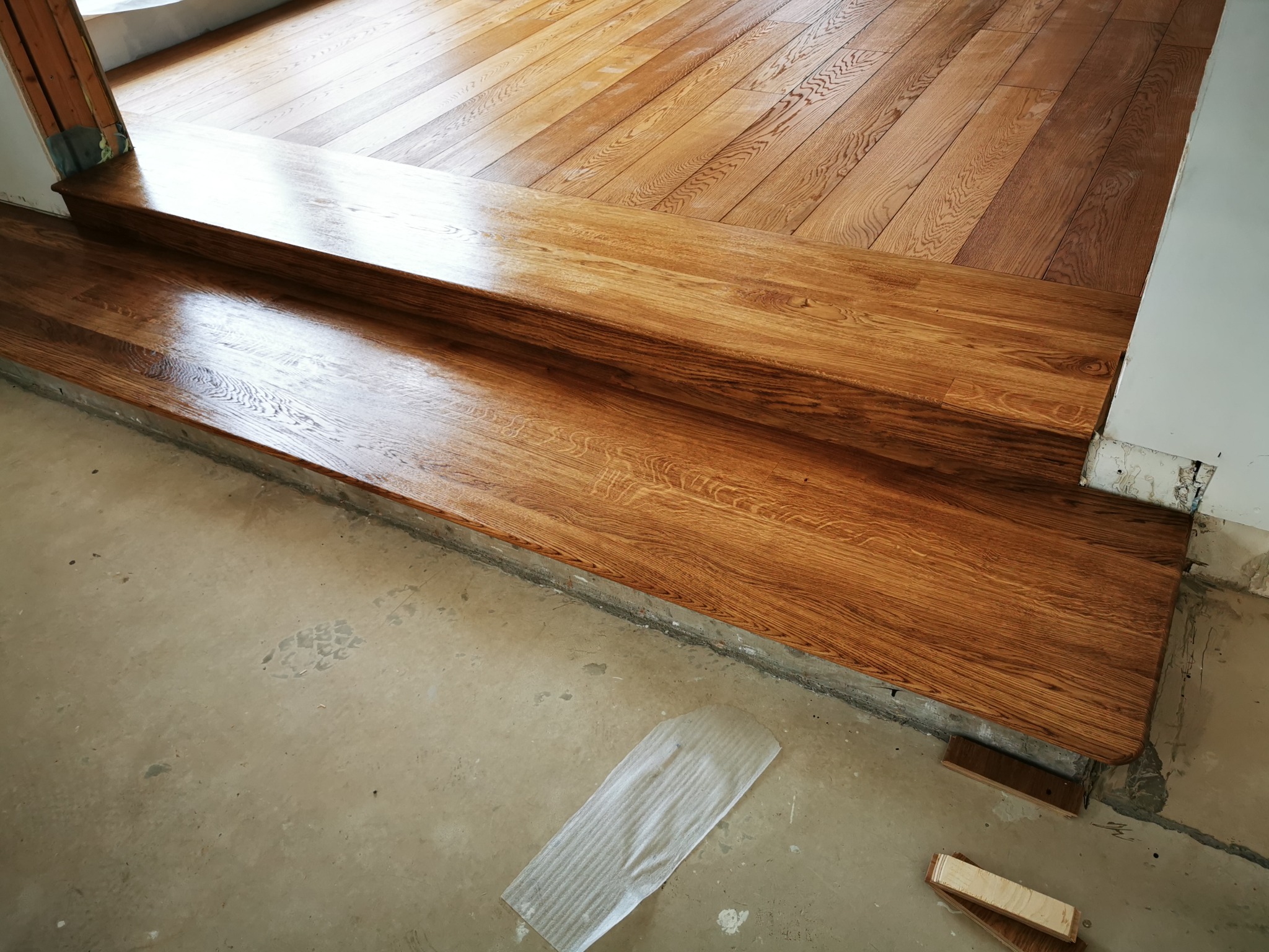 Dwustopniowe schody z desek warstwowych dębowych, widoczne łączenia z podłogą, surowy beton na podłodze, przygotowanie do dalszych prac wykończeniowych.