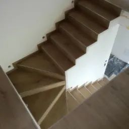 Widok z g&oacute;ry na drewniane schody z oświetleniem punktowym w ścianie, prowadzące w d&oacute;ł, z widocznymi elementami wykończeniowymi i podłogą z paneli.