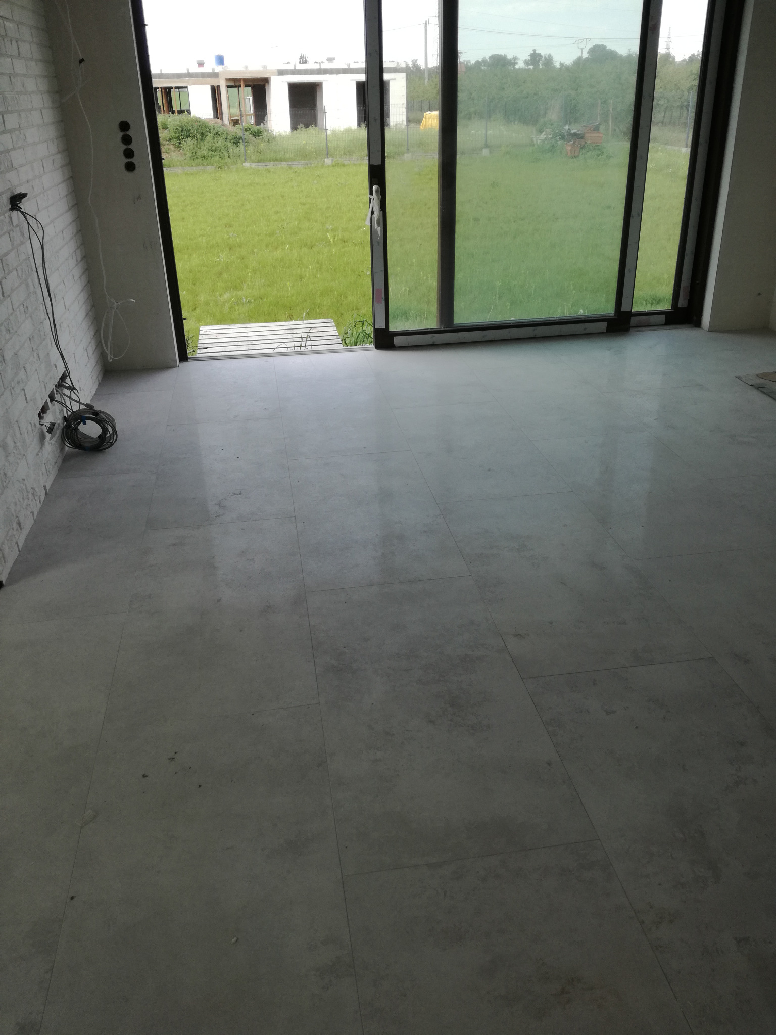 Ułożona szara podłoga z płytek w nowym pomieszczeniu z widokiem na trawnik przez duże okno przesuwne; widoczne kable elektryczne przy ścianie.