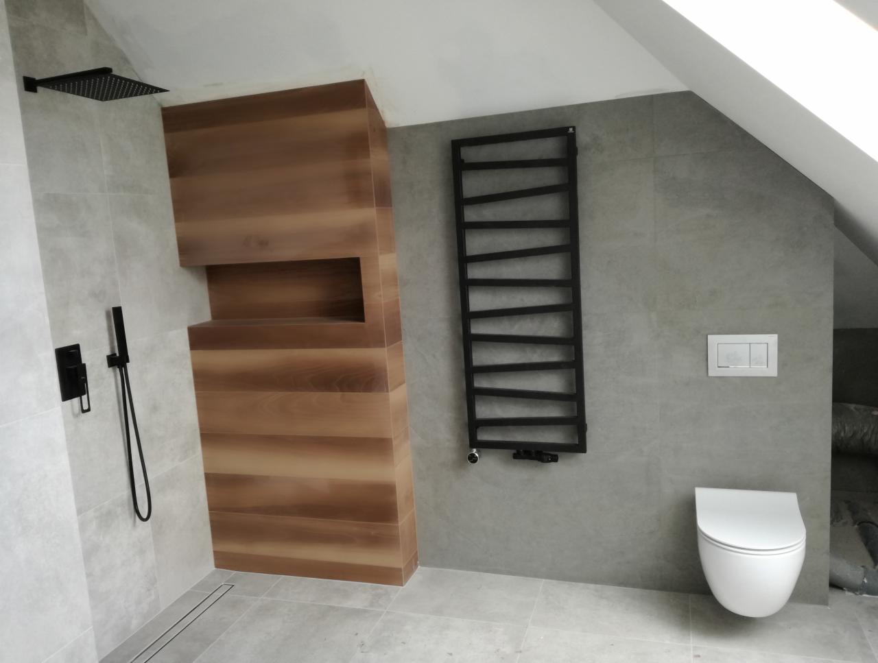 Nowoczesna łazienka na poddaszu z szarymi płytkami, drewnianą zabudową prysznica z wnęką, czarną deszczownicą i grzejnikiem drabinkowym oraz podwieszaną toaletą.