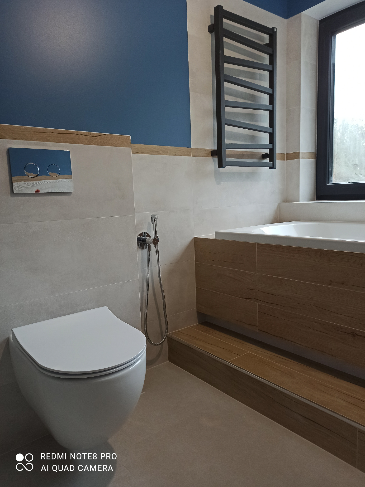 Nowoczesna łazienka z podwieszaną toaletą, bidetką, grzejnikiem drabinkowym w kolorze czarnym, obudowaną wanną z drewnopodobnymi płytkami i niebieską ścianą.