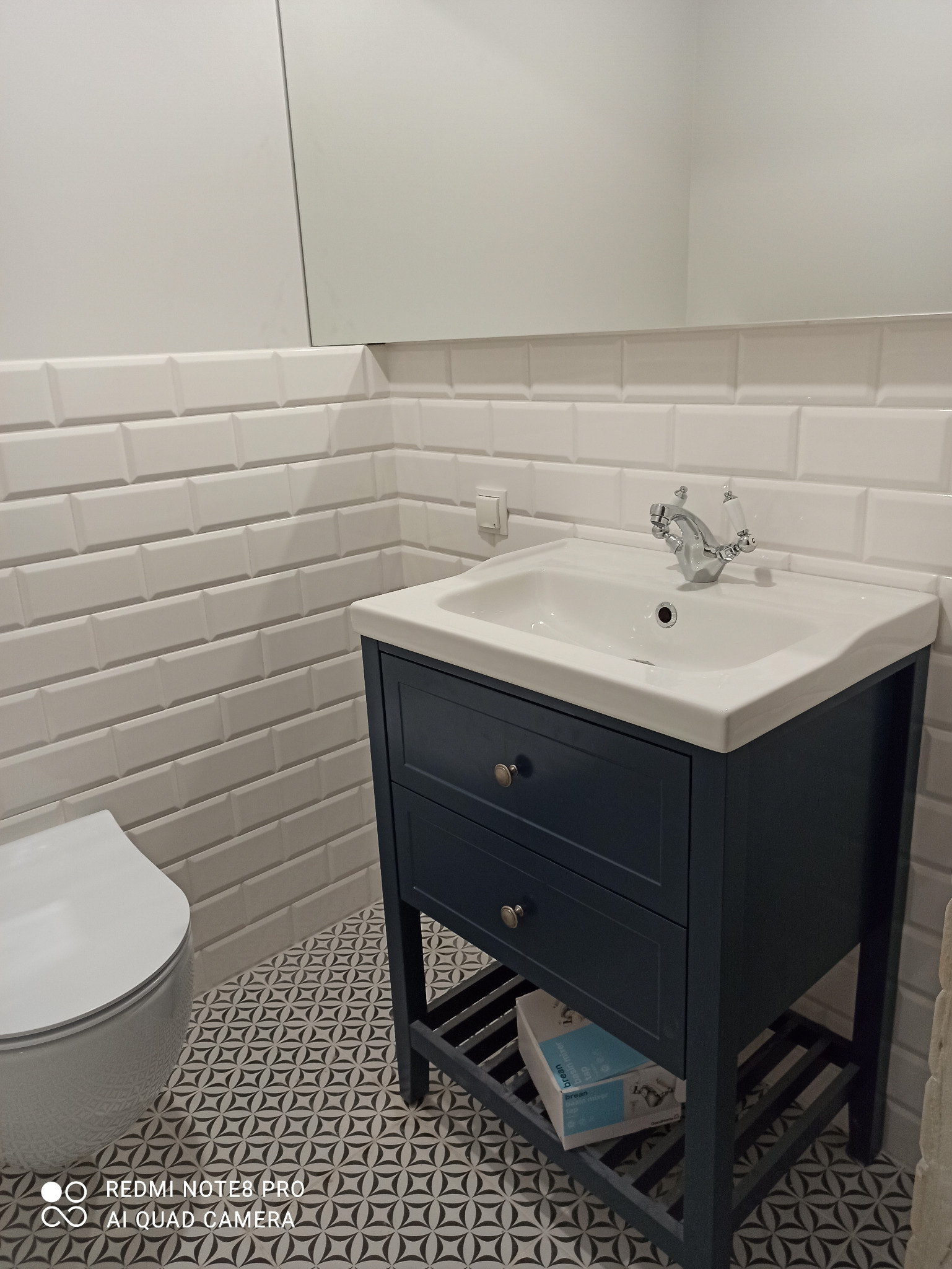 Wykończona łazienka z umywalką w szafce w kolorze granatowym, białe kafelki typu metro i wzorzysta podłoga, widoczny fragment toalety.