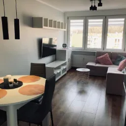 Jasne, przestronne wnętrze salonu z drewnianą podłogą, okrągłym stołem jadalnym z dekoracyjnymi podkładkami i minimalistycznymi czarnymi lampami wiszącymi, w tle sofa z poduszkami w odcieniach różu...