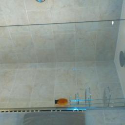 Poziom - Wykończona kabina prysznicowa z beżowymi płytkami imitującymi marmur, szklaną ścianką, chromowanym relingiem na kosmetyki i grzejnikiem ręcznikowym z szarym ręcznikiem.