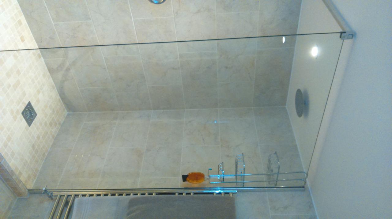 Wykończona kabina prysznicowa z beżowymi płytkami imitującymi marmur, szklaną ścianką, chromowanym relingiem na kosmetyki i grzejnikiem ręcznikowym z szarym ręcznikiem.