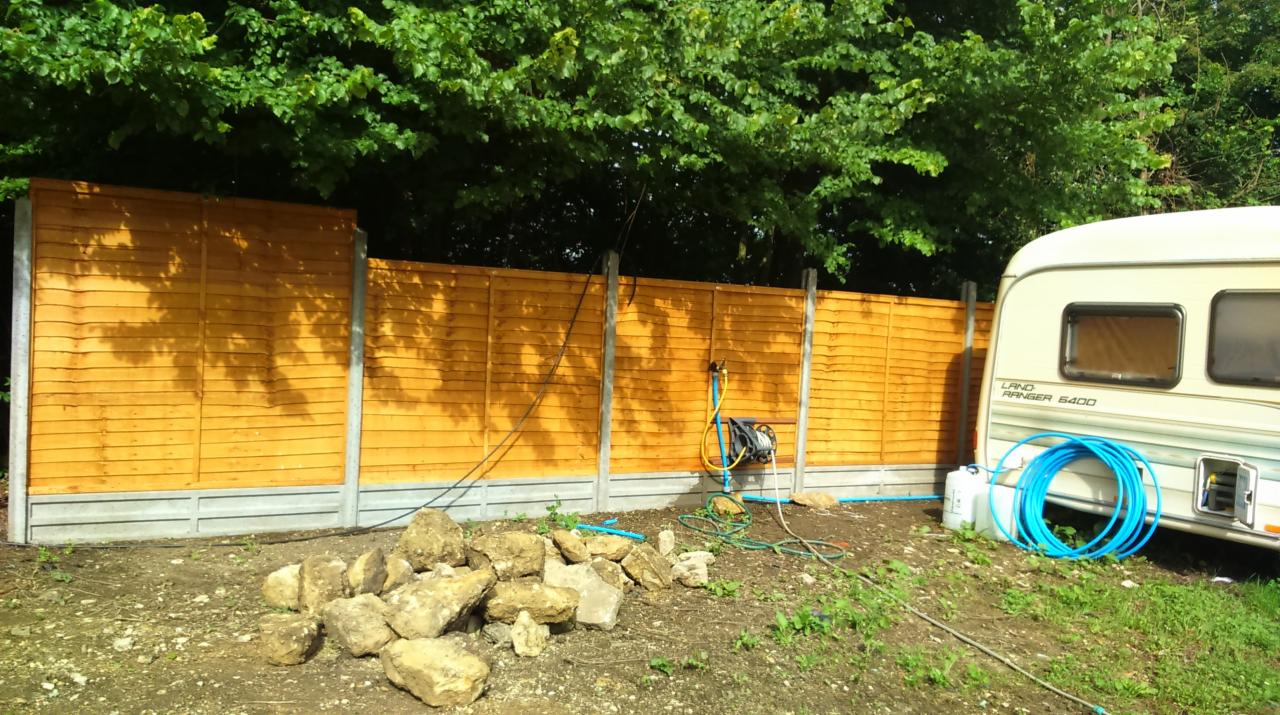 Nowo postawione, drewniane ogrodzenie panelowe z betonową podmurówką, widoczne z boku przyczepy kempingowej, z stertą kamieni na pierwszym planie i zwiniętym wężem ogrodowym oraz myjką ciśnieniową...