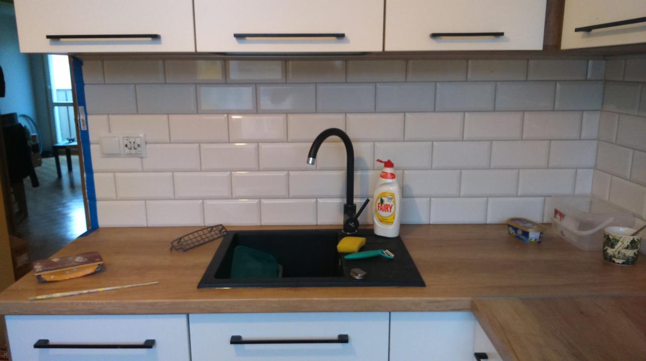 Białe szafki kuchenne z czarnymi uchwytami i czarnym kranem nad czarnym zlewem, białe płytki imitujące cegłę na ścianie, butelka płynu do naczyń Fairy, akcesoria kuchenne na blacie z drewnianym...