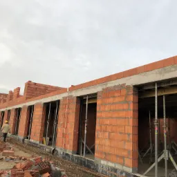 Budowa domu z pustak&oacute;w ceramicznych: widok na wznoszone ściany z otworami okiennymi, podparte metalowymi stemplami, z widocznym pracownikiem w oddali i stertą pustak&oacute;w na pierwszym planie.