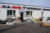 Fasada budynku z napisem 'M.B. SMAK MATERIAŁY BUDOWLANE' nad wejściem, przed budynkiem zgromadzone różnorodne materiały budowlane, widoczne okna na elewacji.