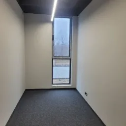 Wnętrze pustego biura w Lubiczu Górnym po malowaniu ścian na biało, z szarą wykładziną podłogową i wąskim oknem z widokiem na zewnątrz.