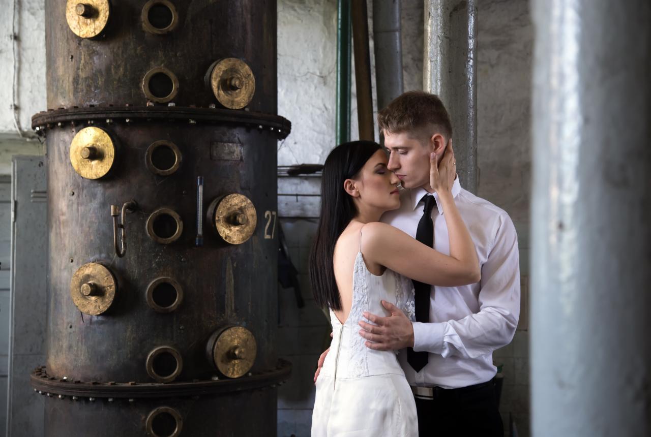 Para w objęciach, panna młoda w białej sukni z odkrytymi plecami, pan młody w białej koszuli i czarnym krawacie, na tle industrialnego zbiornika z metalowymi elementami.
