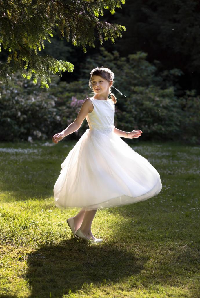 Dziewczynka w białej sukience wiruje na trawie w promieniach słońca, z drzewem w tle.