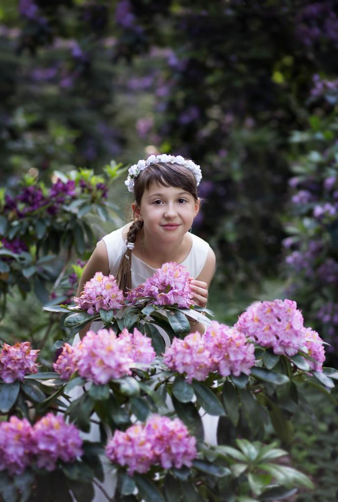 Uśmiechnięta dziewczynka w białej sukience i wianku na głowie, pozuje za krzewem kwitnących różowych rododendronów w ogrodzie.