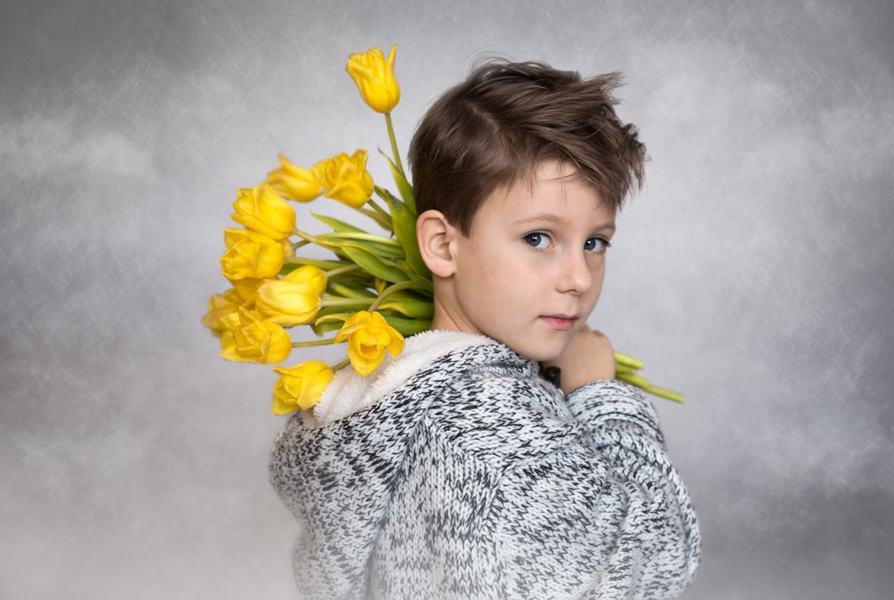 Portret chłopca z niebieskimi oczami trzymającego bukiet żółtych tulipanów na ramieniu, ubrany w sweter z kapturem na rozmytym szarym tle.