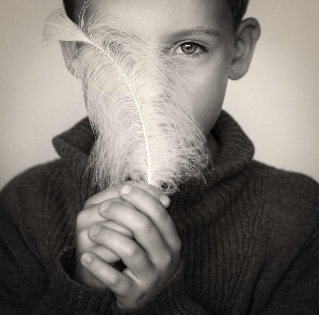 Czarno-białe portretowe ujęcie chłopca, którego część twarzy zasłania delikatne, białe pióro trzymane w złożonych dłoniach, wzrok skierowany w obiektyw.