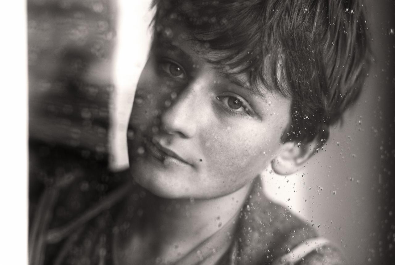 Portret młodego mężczyzny w sepii, widzianego przez zaparowaną szybę z kroplami deszczu, skupiony wzrok w lewo, delikatny uśmiech.