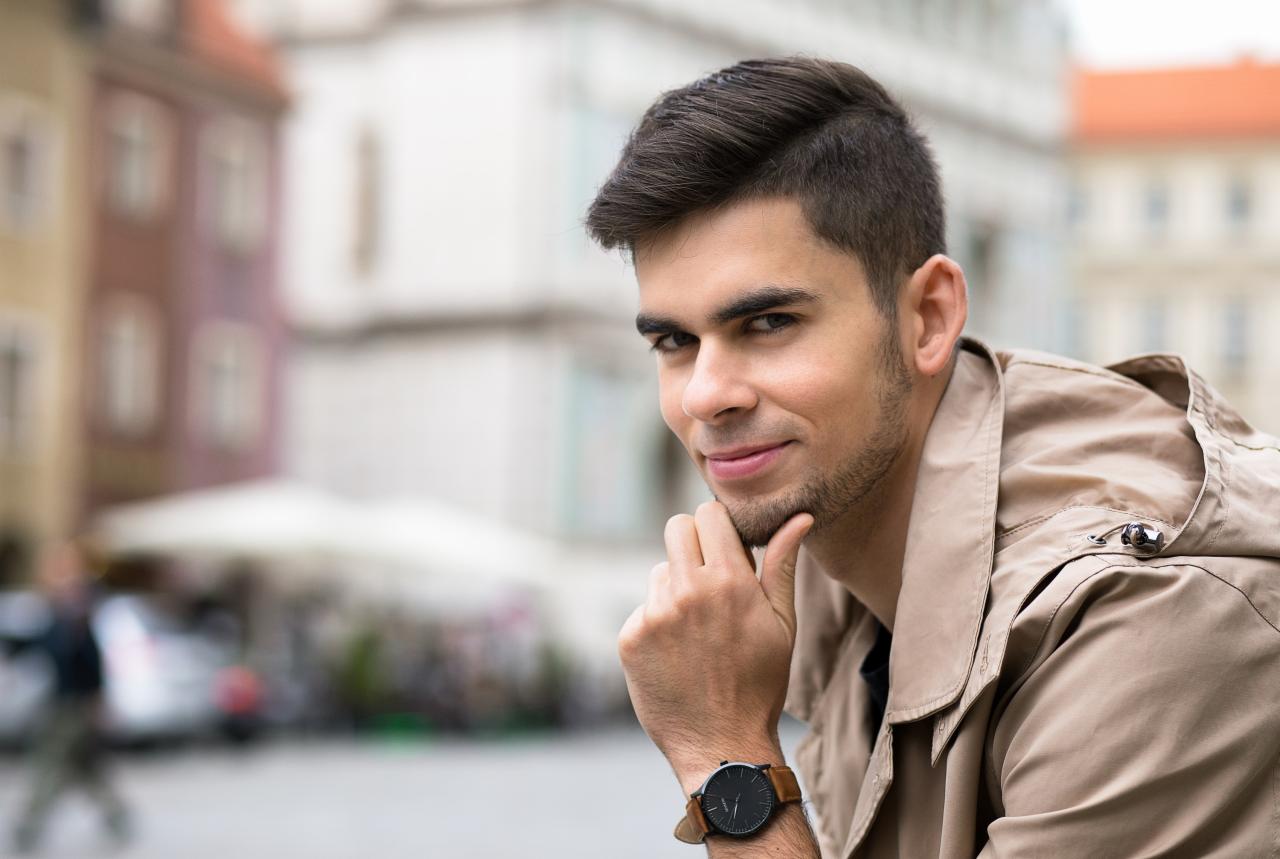 Portret młodego mężczyzny z lekkim zarostem, w beżowej kurtce z kapturem i zegarkiem na skórzanym pasku, pozującego na tle miejskiego placu z rozmytymi budynkami.