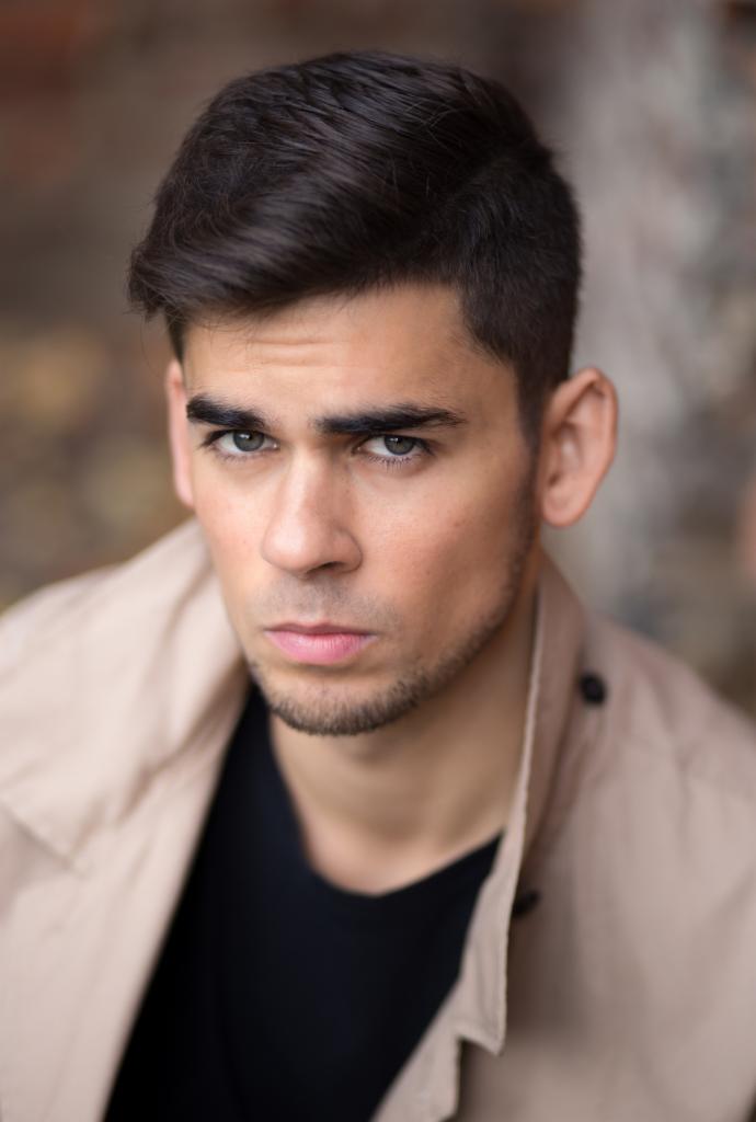 Portret młodego mężczyzny z krótkimi, ciemnymi włosami, w beżowym płaszczu i czarnej koszulce, z intensywnym spojrzeniem skierowanym wprost, na tle rozmytej, ceglanej ściany.