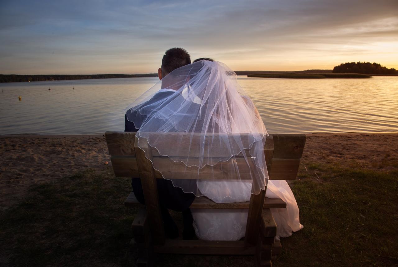 Para młoda siedzi na ławce nad jeziorem o zachodzie słońca, welon panny młodej opada na oparcie ławki.