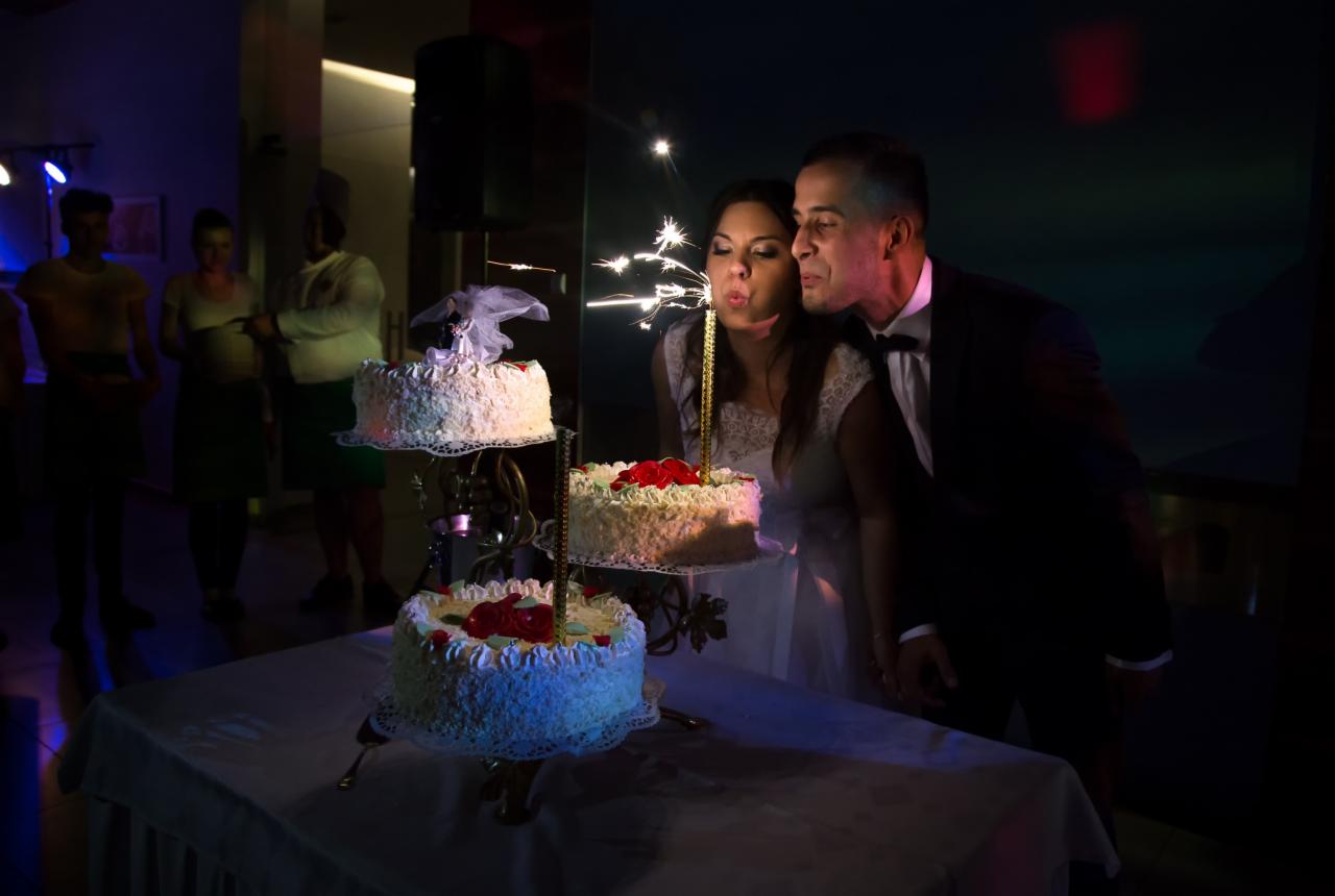 Para nowożeńców w ciemnym otoczeniu zdmuchuje zimne ognie na trzypiętrowym torcie weselnym, w tle widoczni goście.