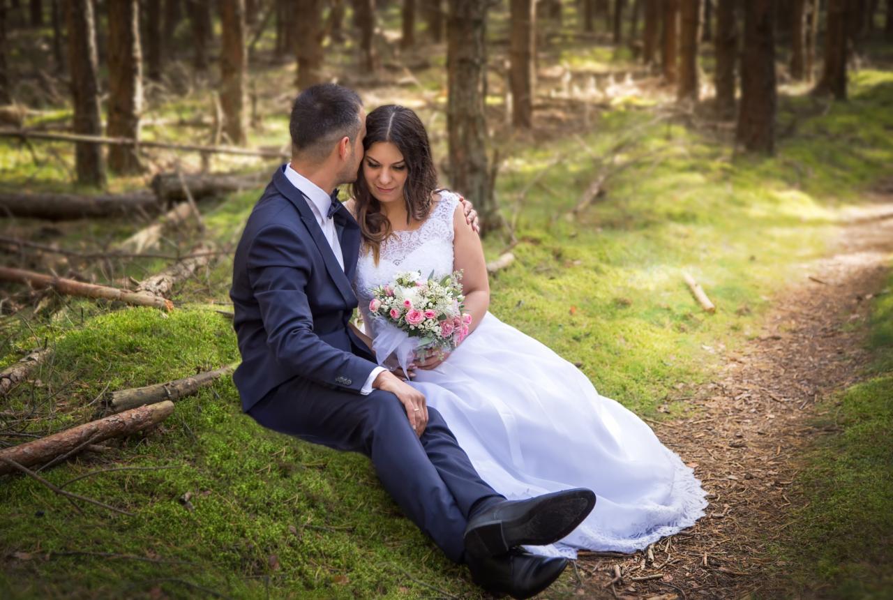 Para młoda siedzi na mchu w lesie, on w granatowym garniturze z muszką, ona w białej sukni z koronkową górą i bukietem różowych i białych kwiatów, delikatnie obejmuje ją.