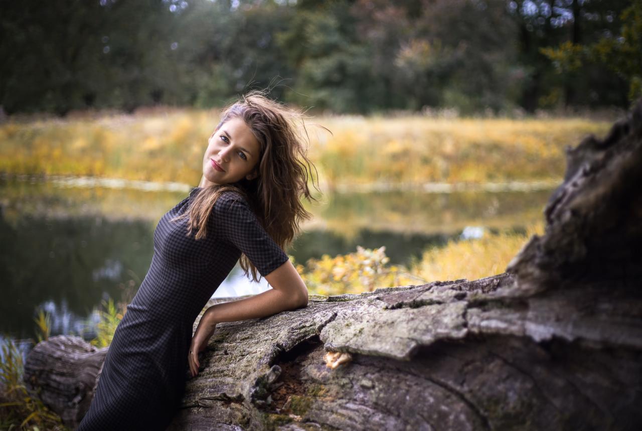 Kobieta w dopasowanej sukience opiera się o spróchniały pień drzewa nad brzegiem jeziora, jesienne tło z zaroślami i odbiciem nieba w wodzie.