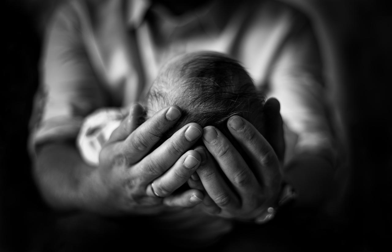 Czarno-białe zbliżenie dłoni dorosłego, delikatnie obejmujących główkę noworodka; widoczny fragment białej koszuli osoby dorosłej, ciemne tło.