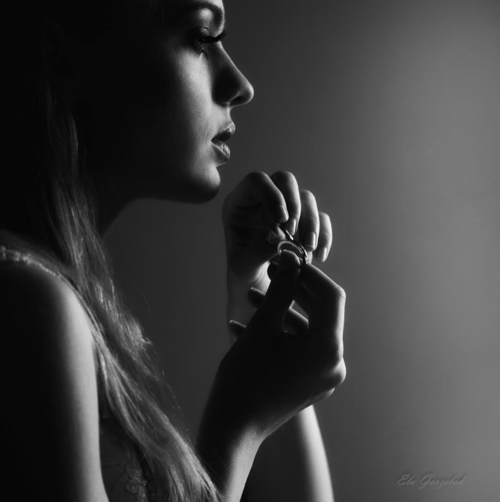 Czarno-białe, profilowe ujęcie kobiety trzymającej w dłoniach pierścionek zaręczynowy, z delikatnym makijażem i długimi rzęsami.