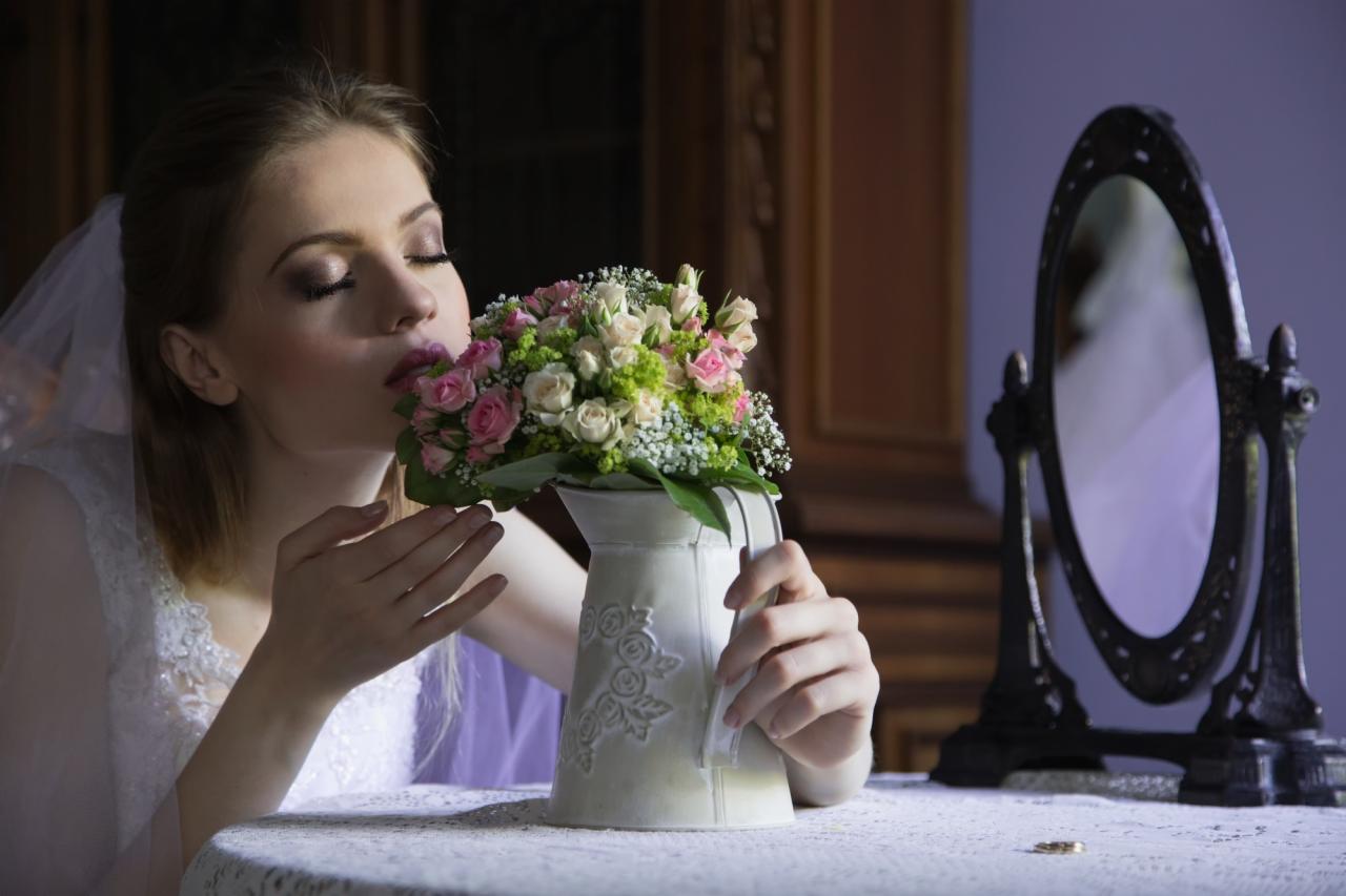 Panna młoda w białej sukni i welonie, z zamkniętymi oczami wącha bukiet różowych i białych róż w ozdobnym dzbanku, na stole leży obrączka, w tle owalne lustro.