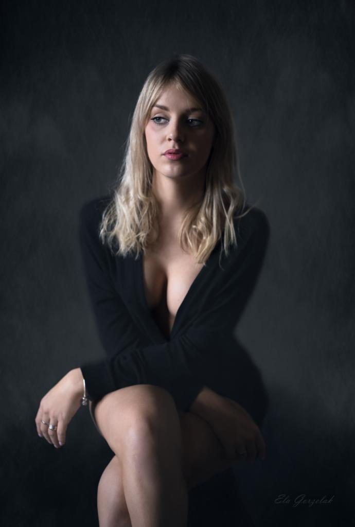 Portret młodej kobiety z blond włosami, siedzącej w czarnej sukience na rozmytym, ciemnoszarym tle. Jej wzrok skierowany jest w lewo, a na dłoniach widoczne są pierścionki i bransoletka.