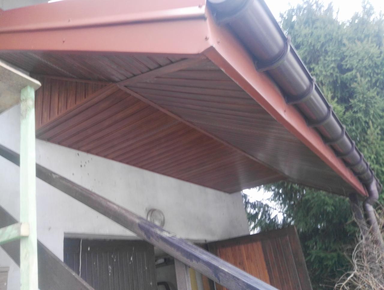 Zadaszenie wejścia do budynku z brązową podbitką i rynnami, widoczne rusztowanie i fragment elewacji.