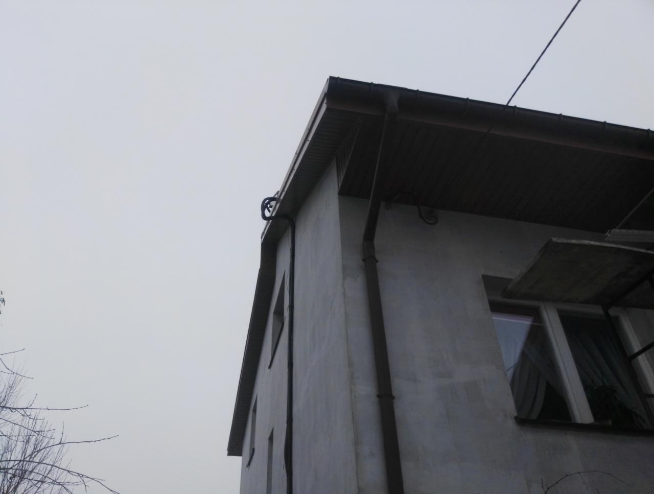 Narożnik budynku z szarą elewacją, brązowym podbitką i rynną, widoczne fragmenty okien i rusztowania, pochmurne niebo w tle.