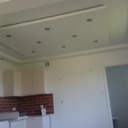 Nowoczesne oświetlenie sufitowe z wbudowanymi punktami świetlnymi w salonie z aneksem kuchennym, widoczna ceglana ściana nad blatem i fragment okna.