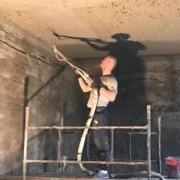 Pracownik na rusztowaniu aplikuje zaprawę na sufit za pomocą agregatu tynkarskiego, widoczny czerwony kubeł i fragment ściany w trakcie obróbki.