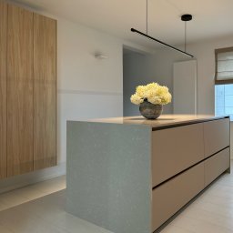 Simple Form Renia Bartoszek - Wnętrze kuchni z wyspą, na której stoi wazon z białymi kwiatami. Nad wyspą zawieszona podłużna lampa. Widoczne drewniane szafki i jasna podłoga.