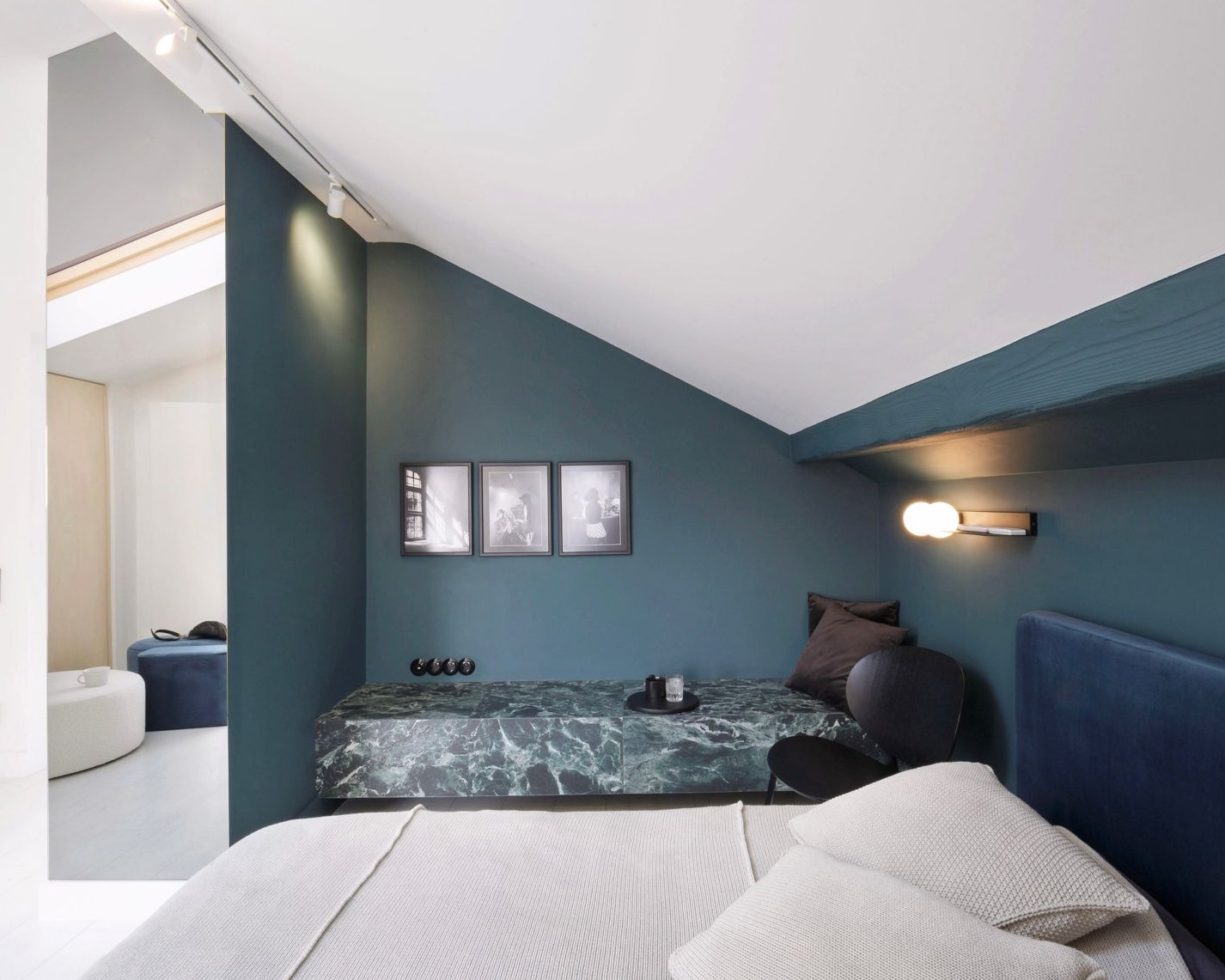 Stylowa sypialnia na poddaszu z zielonymi ścianami i marmurowym siedziskiem. Trzy czarno-białe zdjęcia nad siedziskiem. Minimalistyczna lampka ścienna.