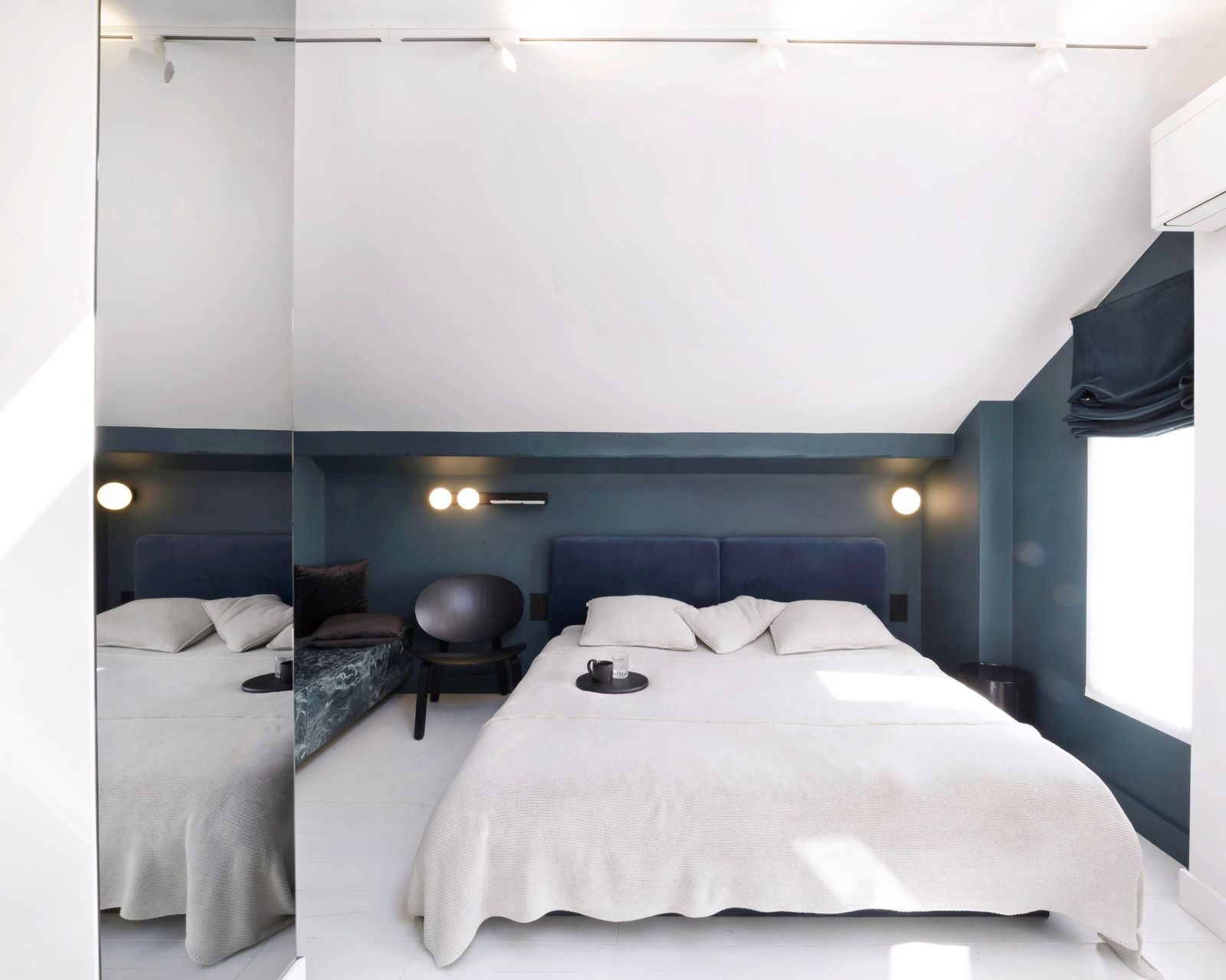 Stylowa sypialnia w odcieniach granatu z dużym lustrem, białymi podłogami i lampami ściennymi. Na łóżku taca z filiżanką i szklanką.