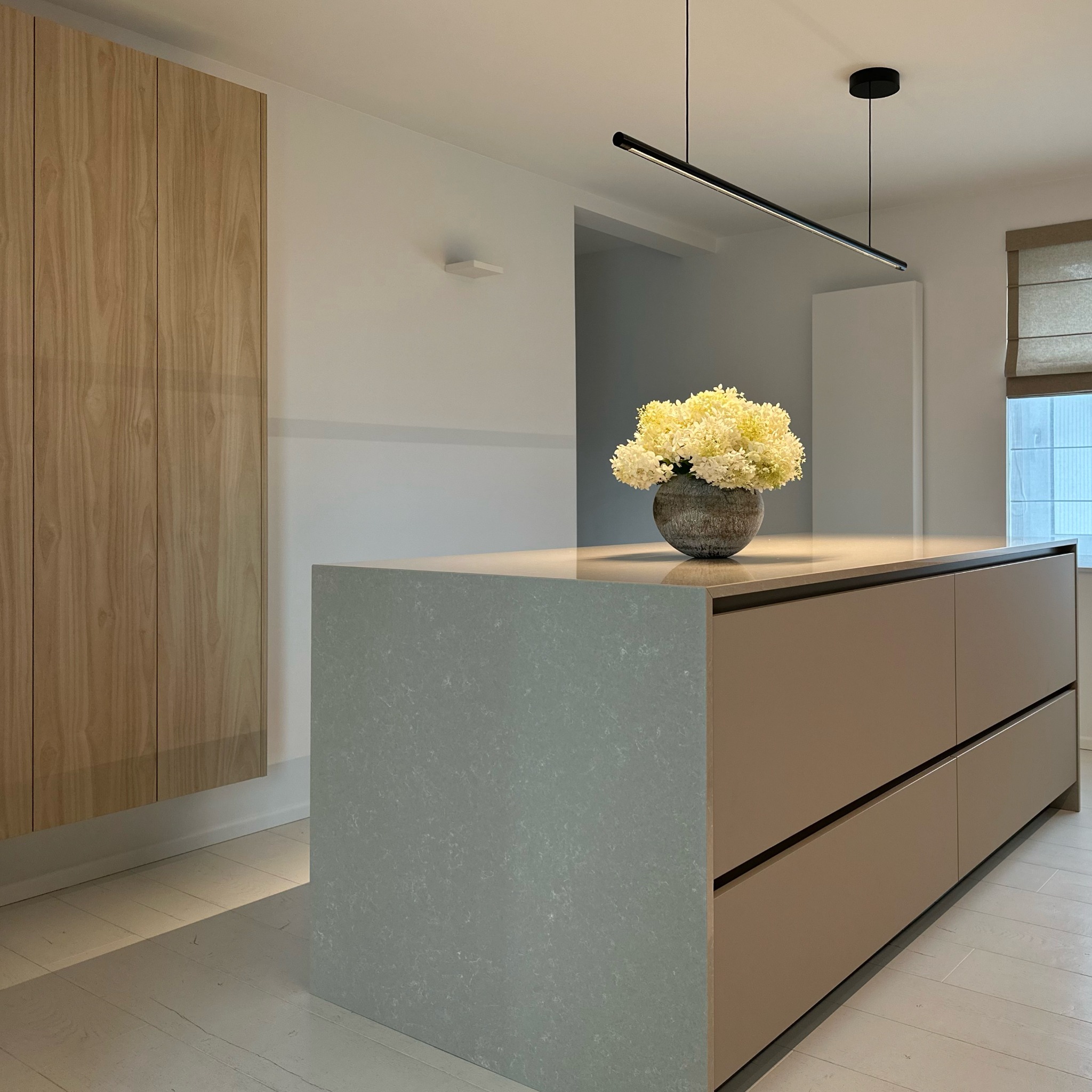 Wnętrze kuchni z wyspą, na której stoi wazon z białymi kwiatami. Nad wyspą zawieszona podłużna lampa. Widoczne drewniane szafki i jasna podłoga.