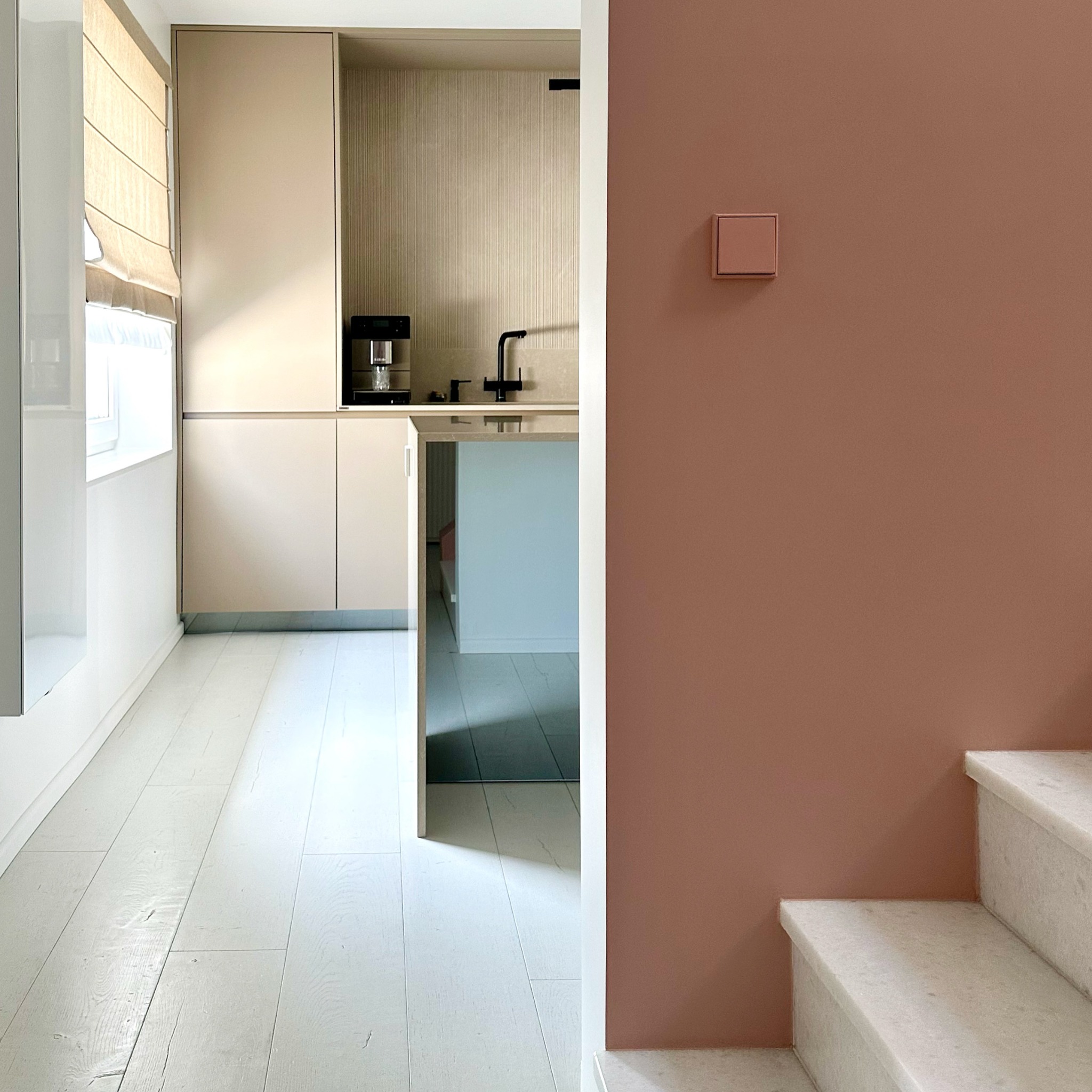 Minimalistyczna kuchnia z beżowymi szafkami, białymi drewnianymi podłogami i ścianą w kolorze dusty rose. Widoczny ekspres do kawy i czarna bateria.