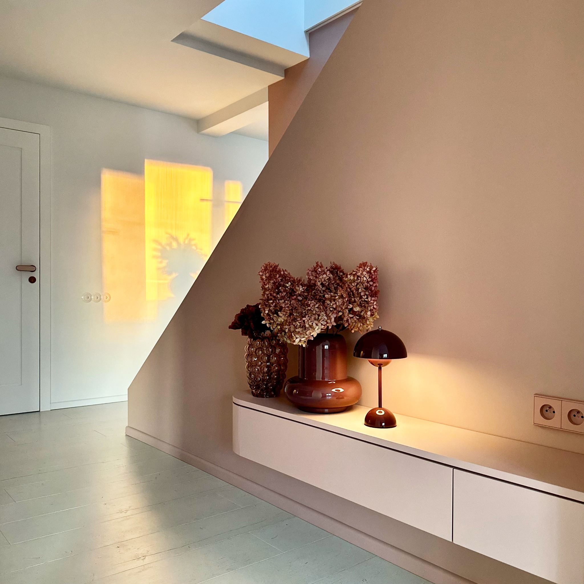 Jasne wnętrze z drewnianą podłogą. Na ścianie wazon z kwiatami, lampka i kontakt. W tle drzwi i okno z odbijającym się światłem. Minimalistyczny design.