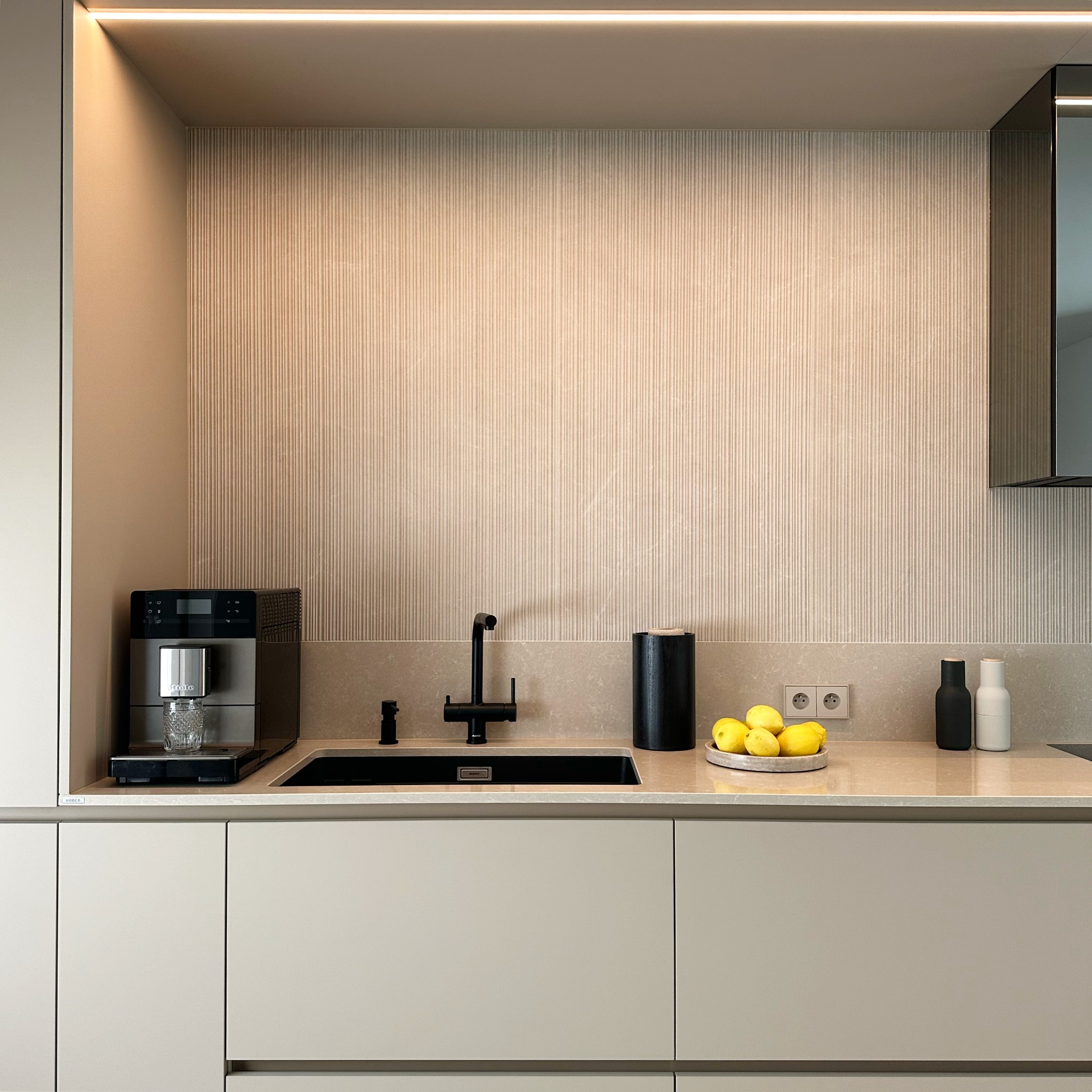 Minimalistyczna kuchnia z beżowymi frontami, czarną armaturą i ekspres do kawy Miele. Ściana z subtelną, pionową fakturą i podświetleniem LED.