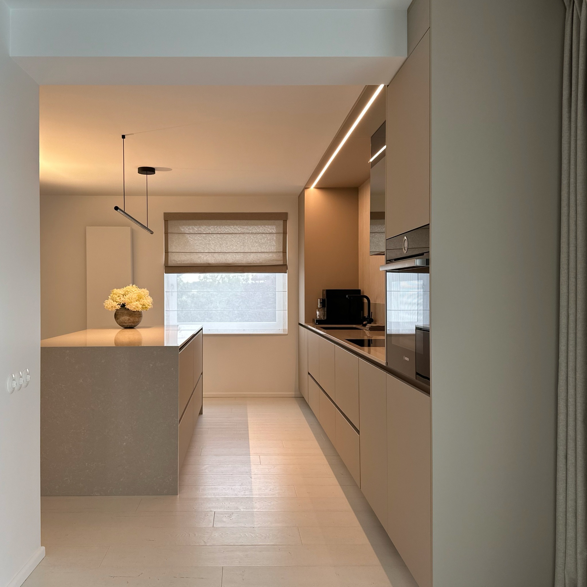 Beżowa, minimalistyczna kuchnia z wyspą i subtelnym oświetleniem LED. Na wyspie wazon z kwiatami, w tle okno z roletą rzymską.