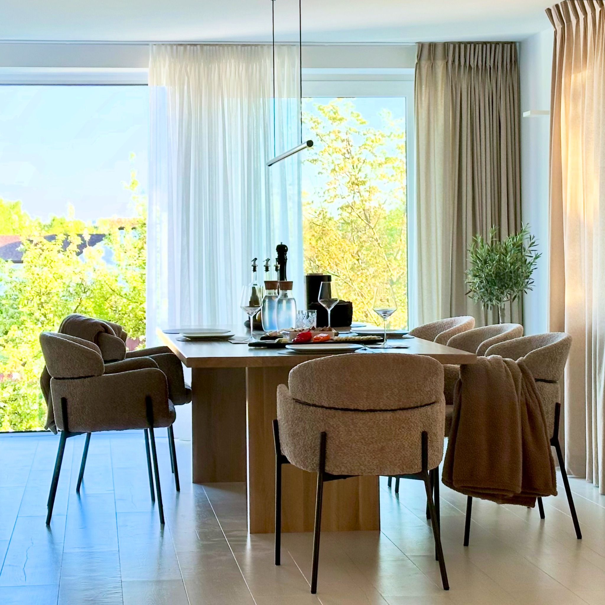 Jasna jadalnia z drewnianym stołem, krzesłami obitymi beżową tkaniną, dużymi oknami z zasłonami i minimalistyczną lampą wiszącą. W tle widok na zieleń.