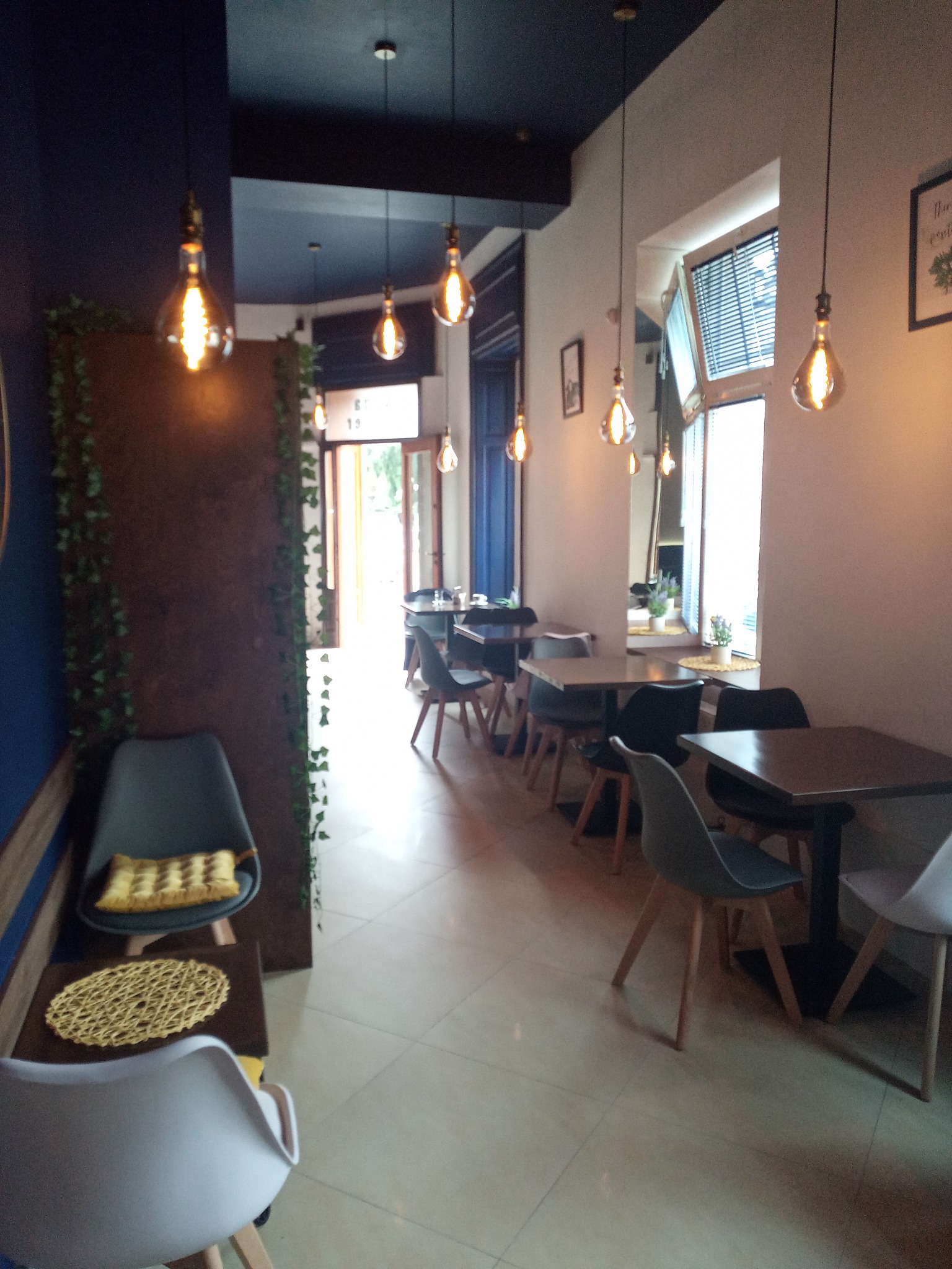 Wnętrze kawiarni z rzędem stolików i krzeseł, oświetlonych wiszącymi żarówkami w stylu retro, dekorowane roślinami pnącymi i elementami w kolorze granatowym.