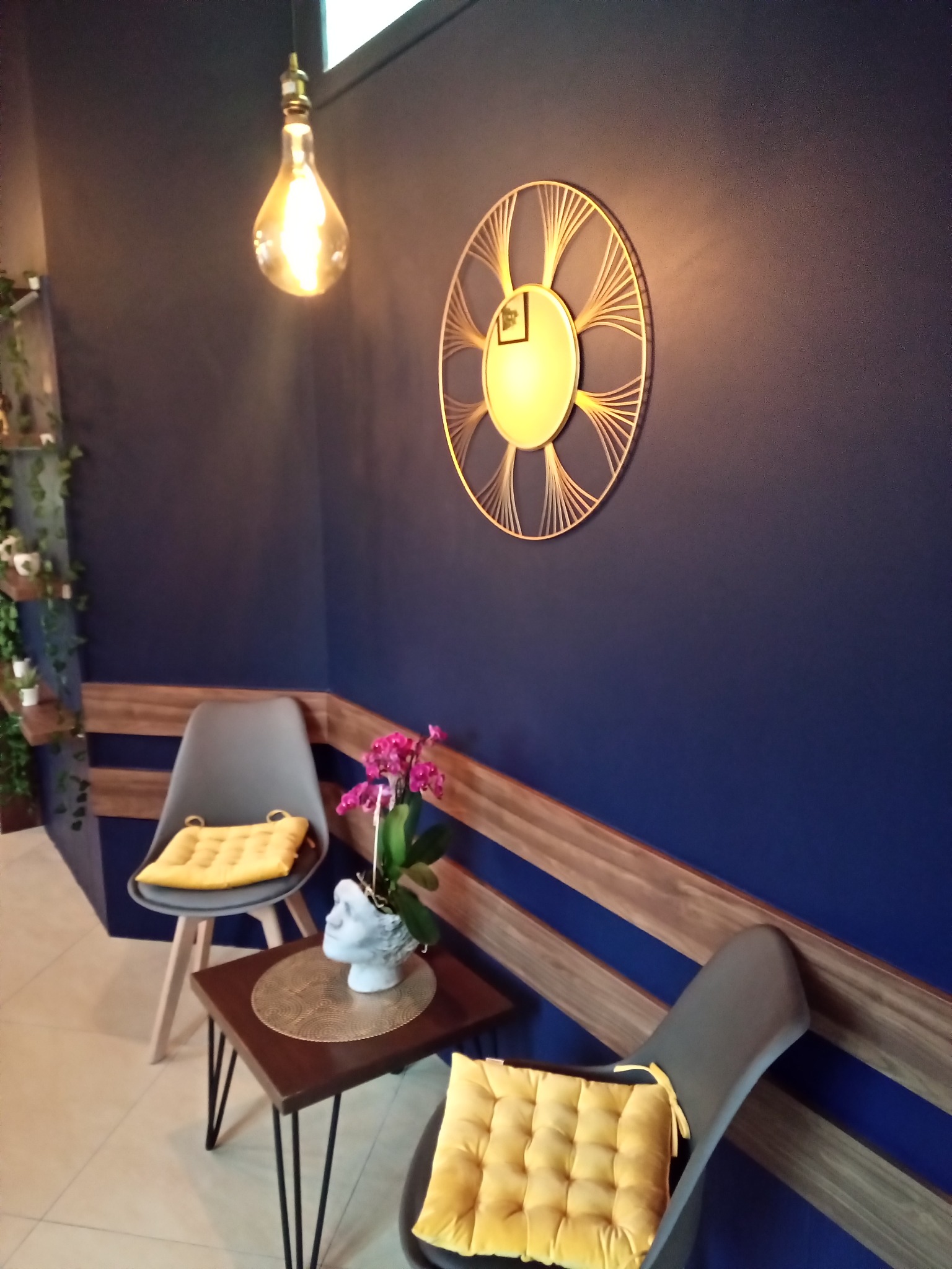 Niebieskie wnętrze z dekoracyjnym lustrem, żarówką Edison, stolikiem z kwiatami w głowie-doniczce i szarymi krzesłami z żółtymi poduszkami.