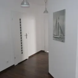 Wnętrze przedpokoju po remoncie: białe ściany, ciemna drewniana podłoga, nowoczesne oświetlenie, drzwi z dekoracyjnymi wgłębieniami i obraz z motywem żaglówki na ścianie.